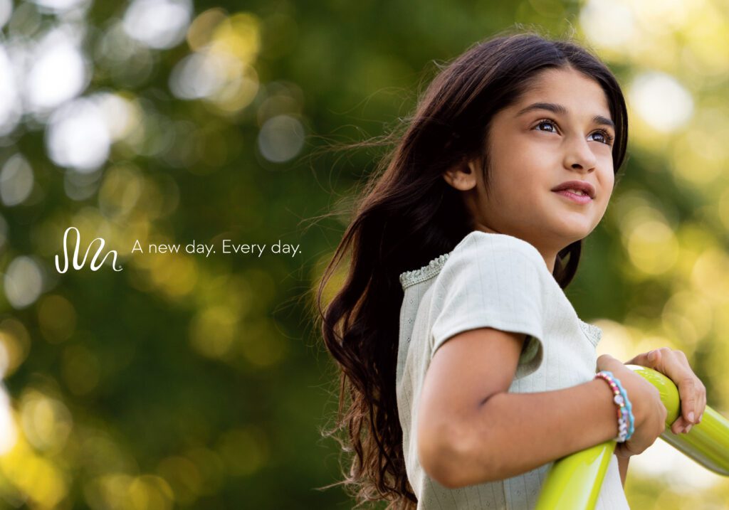 Smiling girl holds yellow bar and looks up, with green foliage behind her. Text: “A new day. Every day.”.