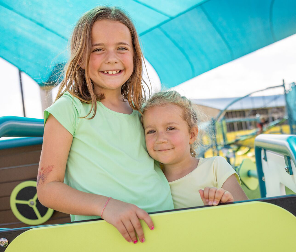 Two young girls smile together at a playground, standing beneath a big blue canopy on a sunny day.