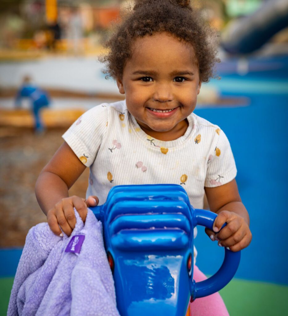 Young child smiles while holding a blue playground toy and purple blanket at an outdoor play area.