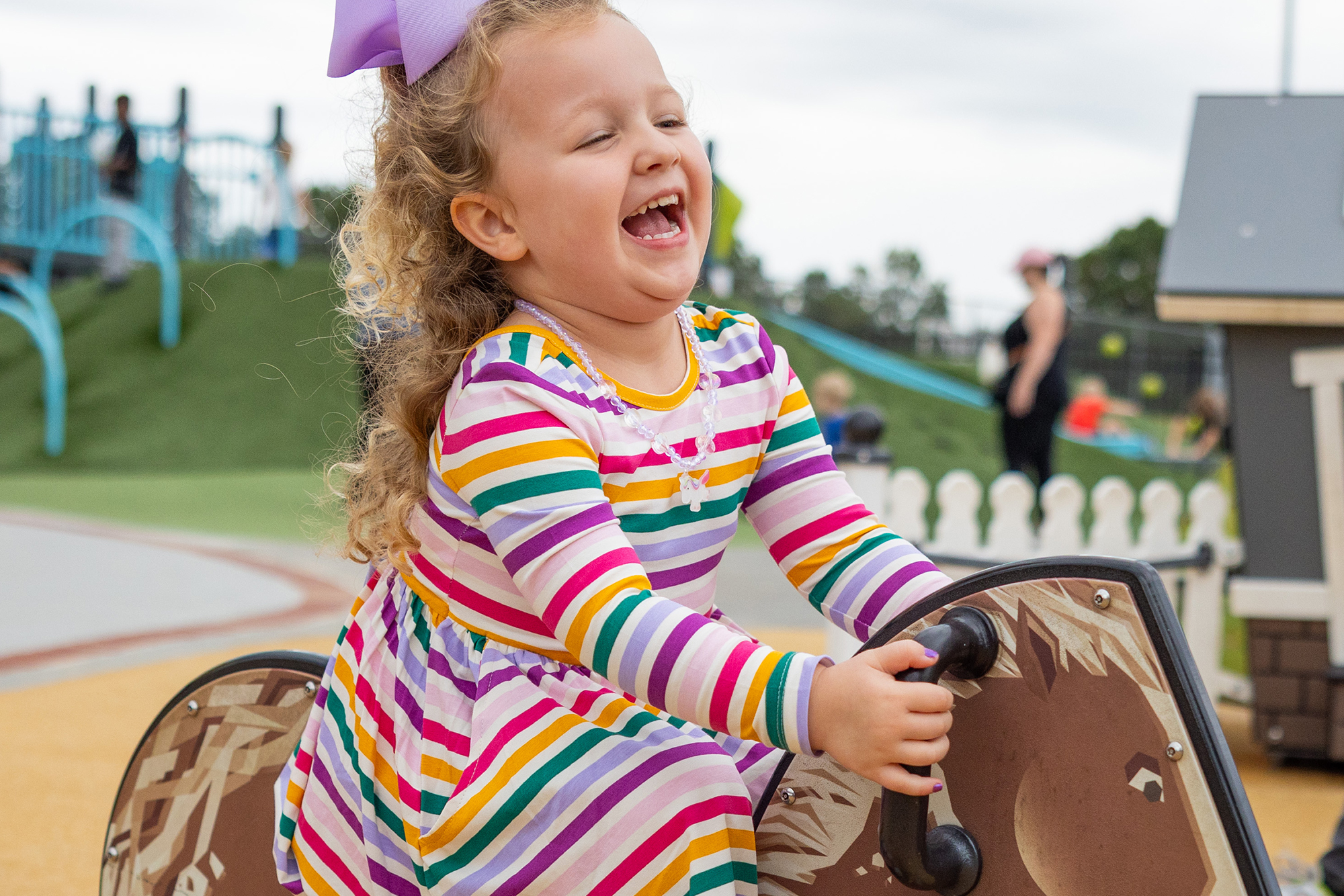 Smiling girl in a striped dress laughs while playing on a playground rocking toy outside.