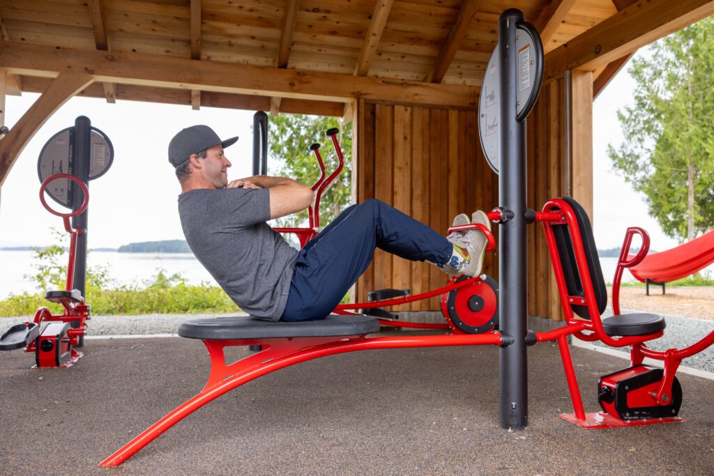 A man does sit-ups on red outdoor gym equipment beneath a wooden shelter beside a lakeshore.