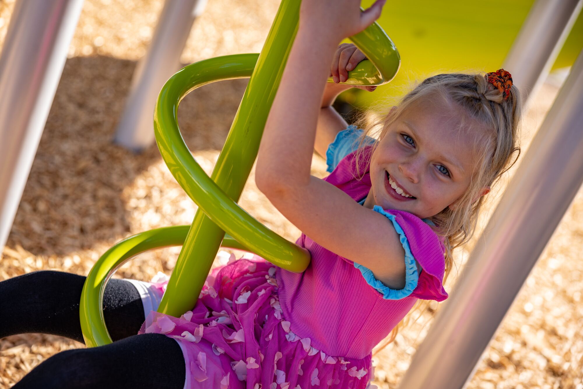 Girl in a pink dress smiles while climbing a green spiral play structure at the playground.