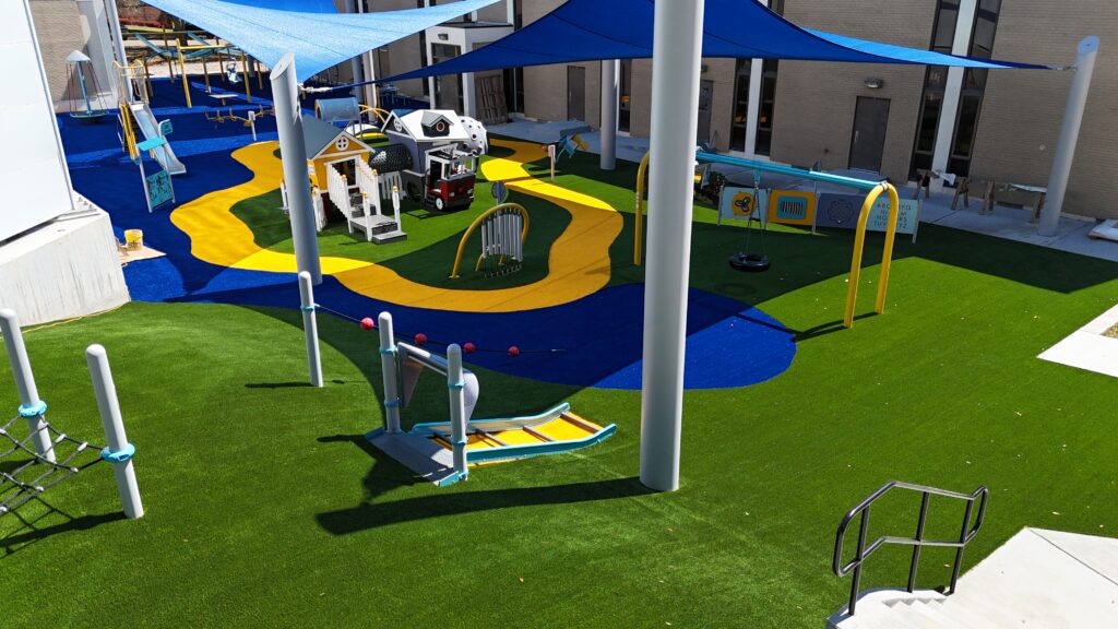 Playground with bright turf, slides, playhouses, and shade structures under a sunny sky.