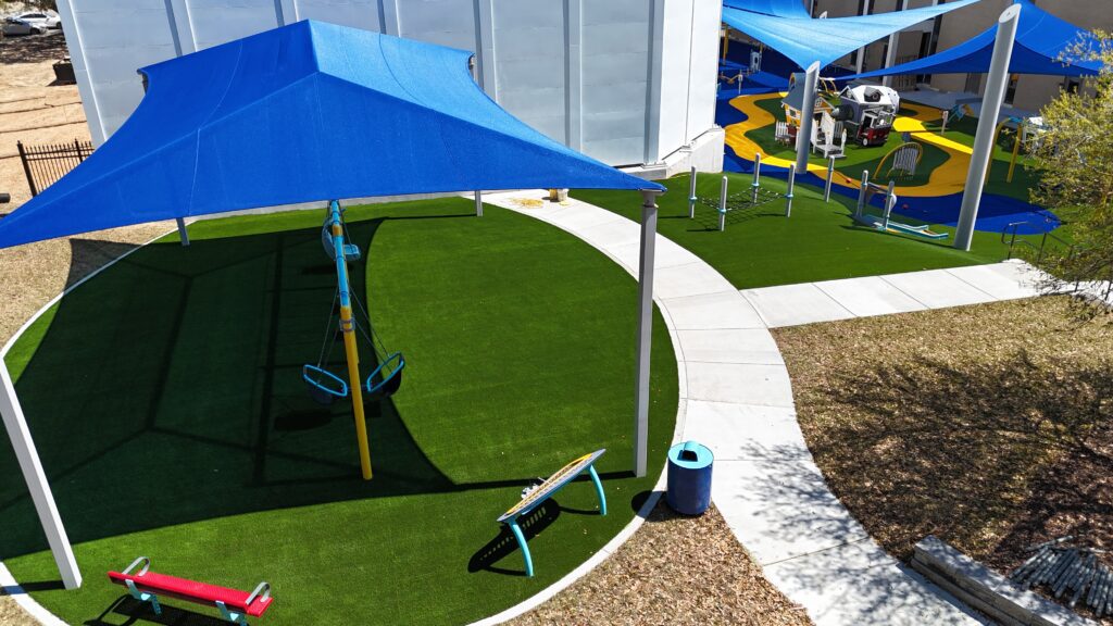 Playground with green turf, blue canopies, benches, and play equipment beside a large white building.