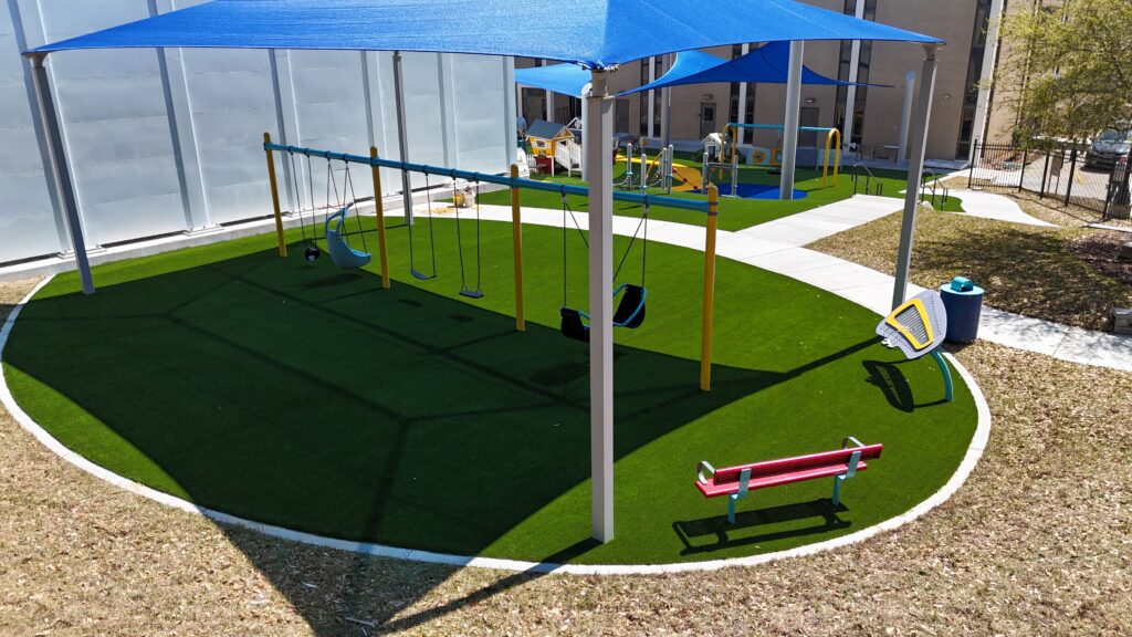 Playground with swings, slides, and benches on artificial grass beneath blue canopies on a sunny day.