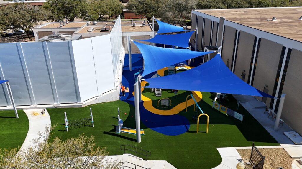 Playground with blue shade sails, yellow walkways, and play equipment on artificial grass between two buildings.