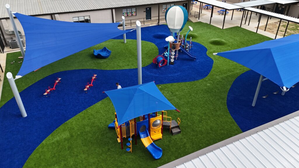 Playground seen from above with blue shade sails, play areas, and winding blue paths across green turf.