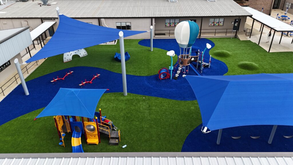 Playground featuring blue shade sails, play equipment, and artificial grass with curving blue pathways.