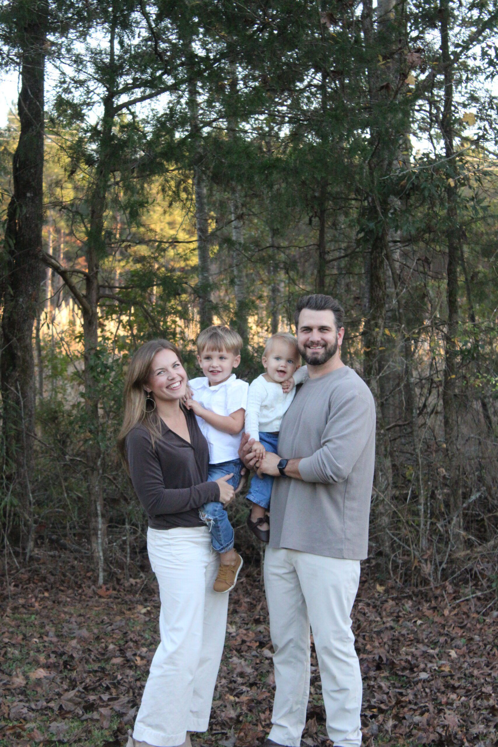 A smiling family of four stands outside near trees, each parent holding a young child.