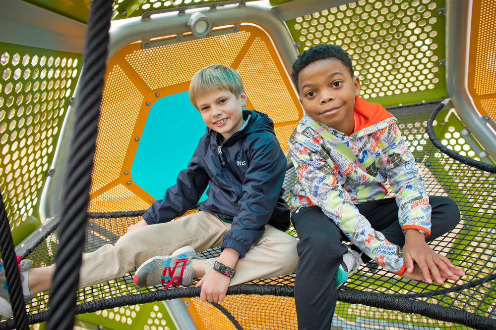 Two boys smile as they sit on a netted play structure with bright, colorful perforated panels in the background.