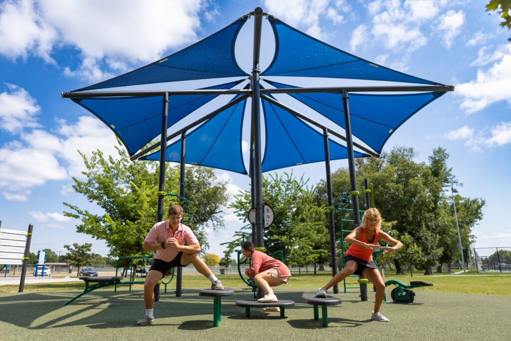 Three people work out beneath a large blue shade canopy at an outdoor fitness park on a sunny day.