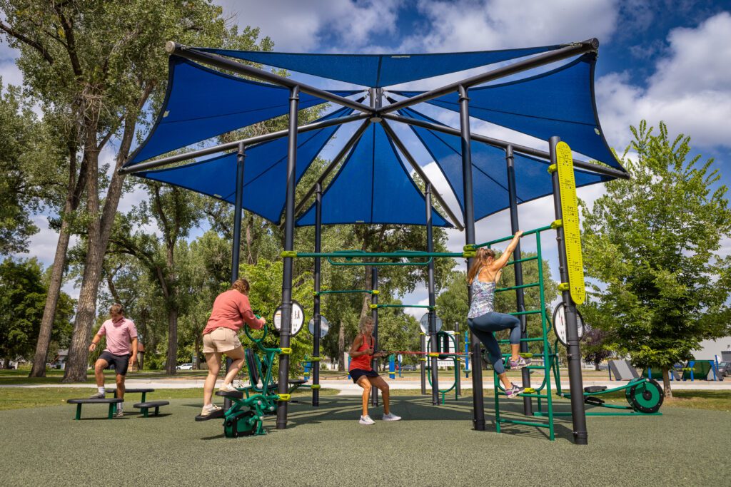 Four people exercise on outdoor equipment beneath a blue canopy in a park on a sunny day.