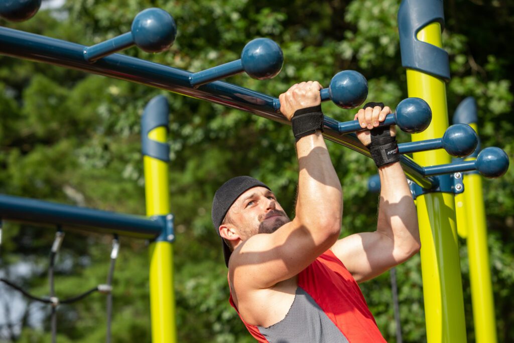Man wearing a red tank top and black headband performs pull-ups on monkey bars at an outdoor gym.