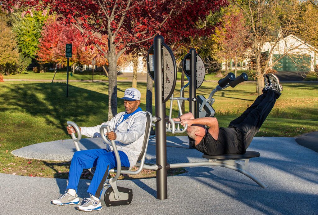 Two men exercise on outdoor gym equipment in a park, surrounded by autumn trees with colorful leaves.