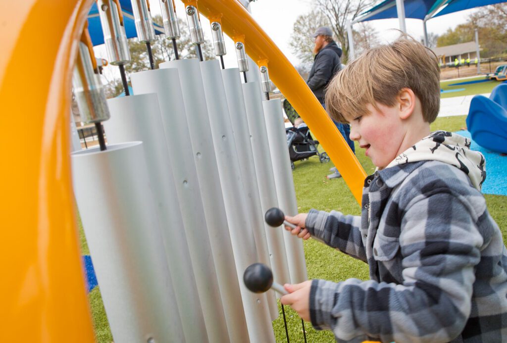 Smiling young boy uses mallets to play large outdoor chimes at a playground, appearing happy and focused.