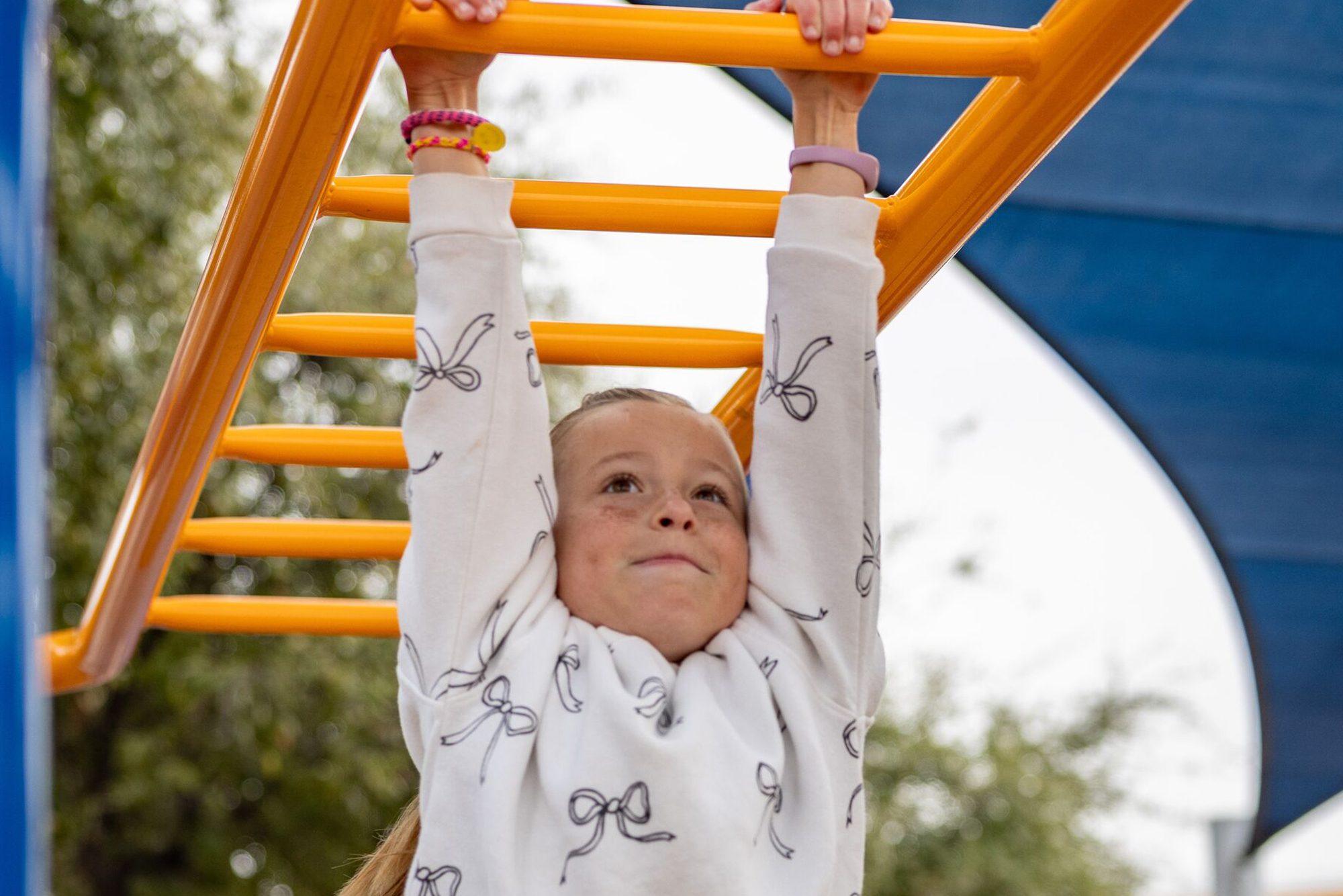 Smiling child in a white sweater hangs from yellow monkey bars at a playground, looking up.