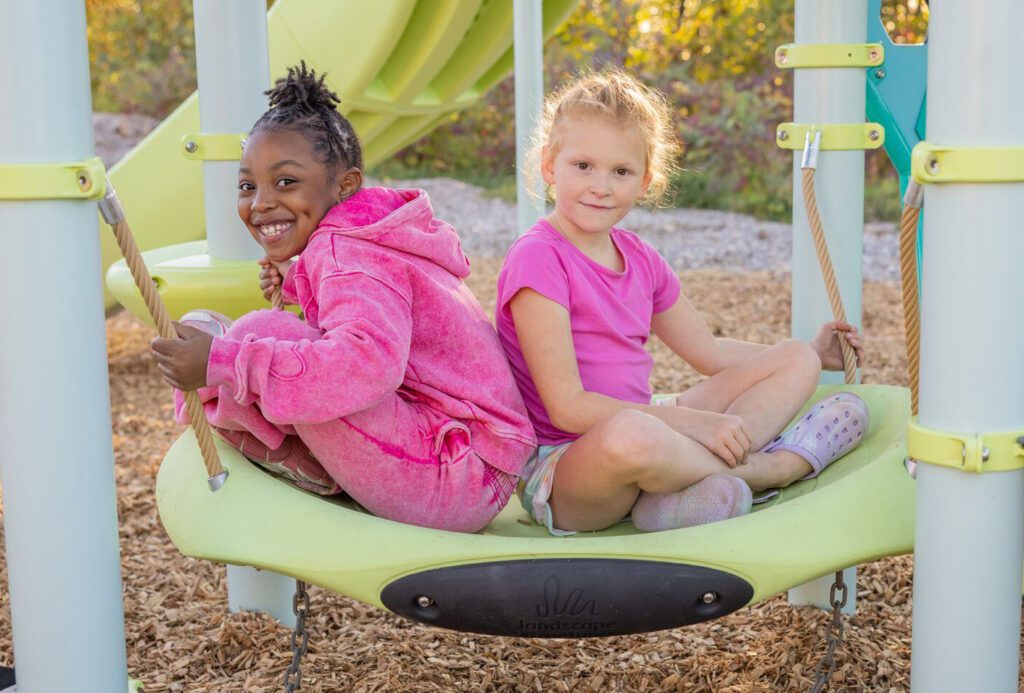 Two young girls sit back-to-back on a playground swing, both smiling happily at the camera.