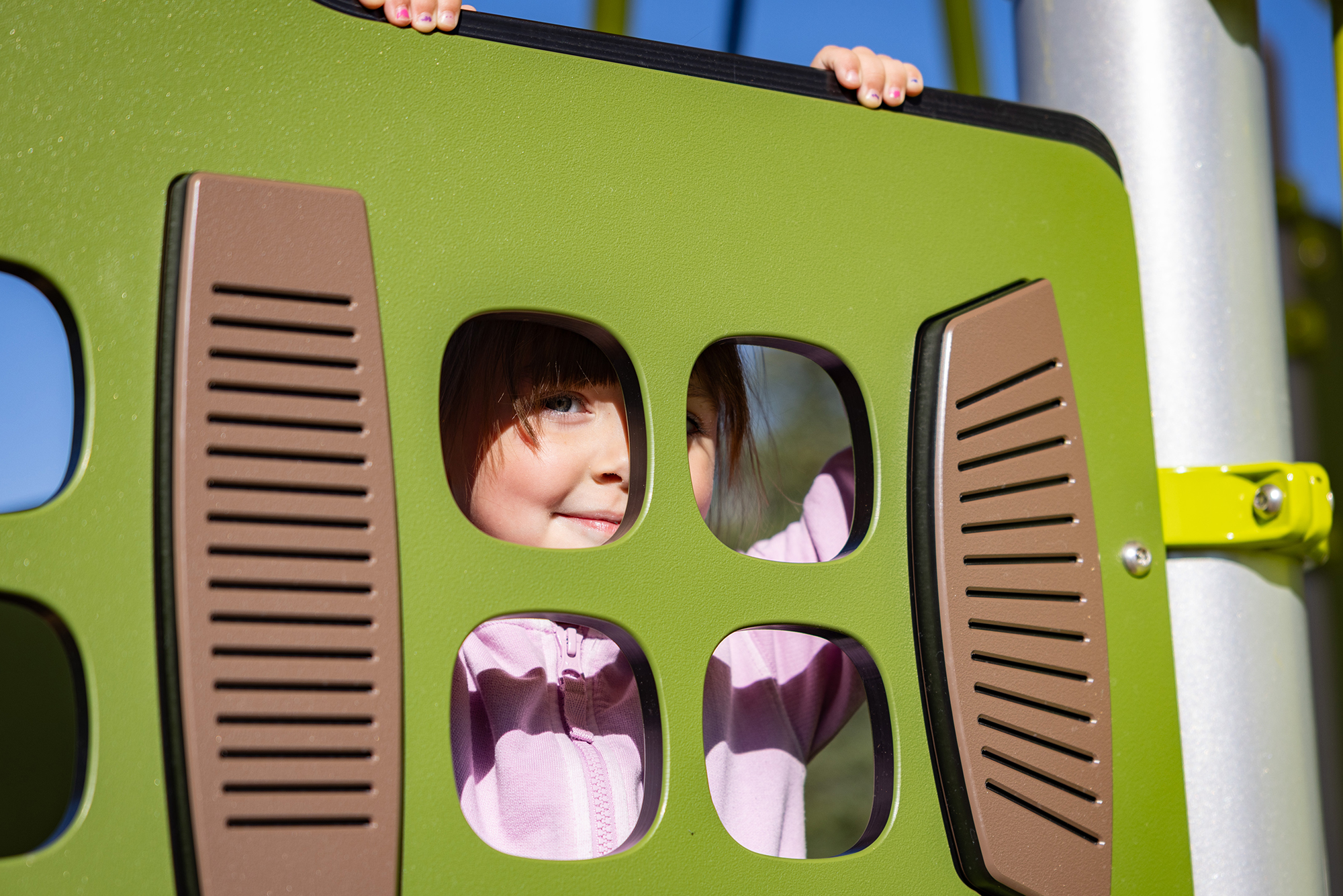Smiling child in a pink jacket peeks through holes in bright green playground equipment.