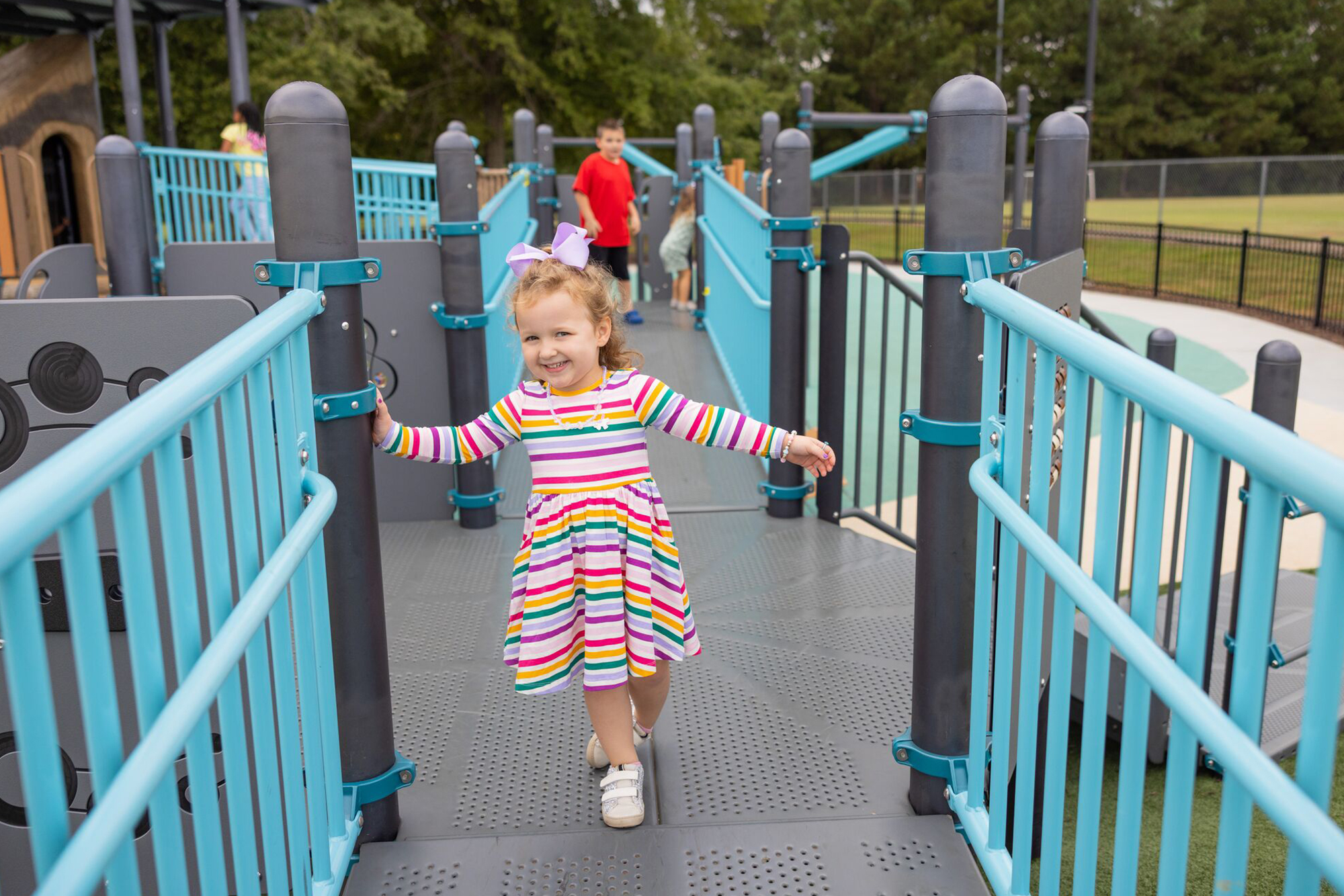 Young girl in a bright striped dress walks on a blue playground bridge, other children play in the background.