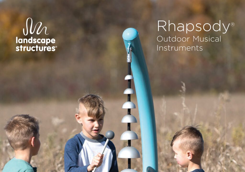 Three boys strike outdoor musical bells with mallets beneath a "Rhapsody Outdoor Musical Instruments" sign in a field.