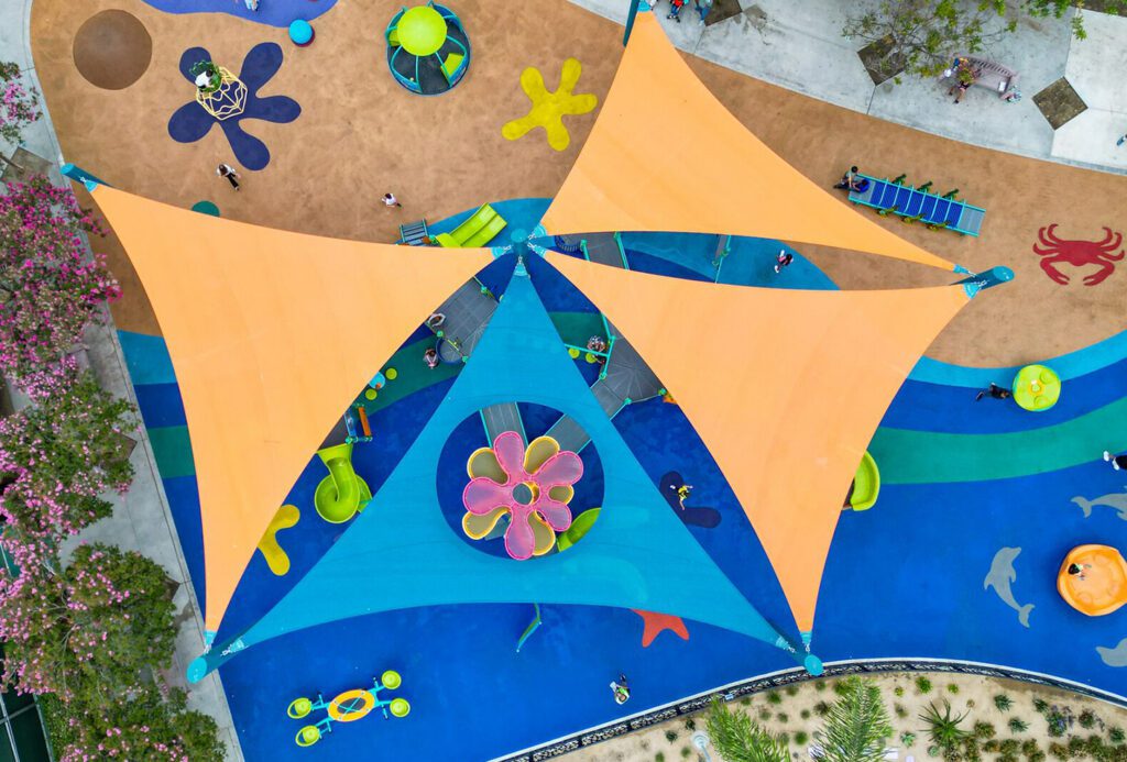 Aerial view of a vibrant playground with orange and blue shade sails covering multiple play structures.