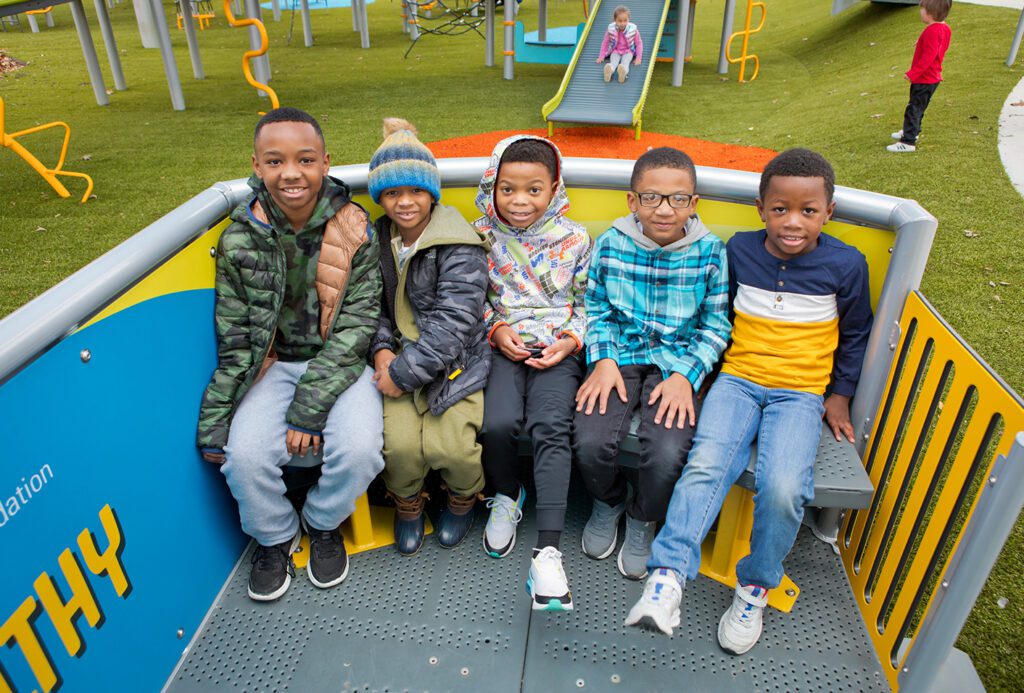Five kids sit on playground equipment, smiling at the camera with slides and grassy area in the background.