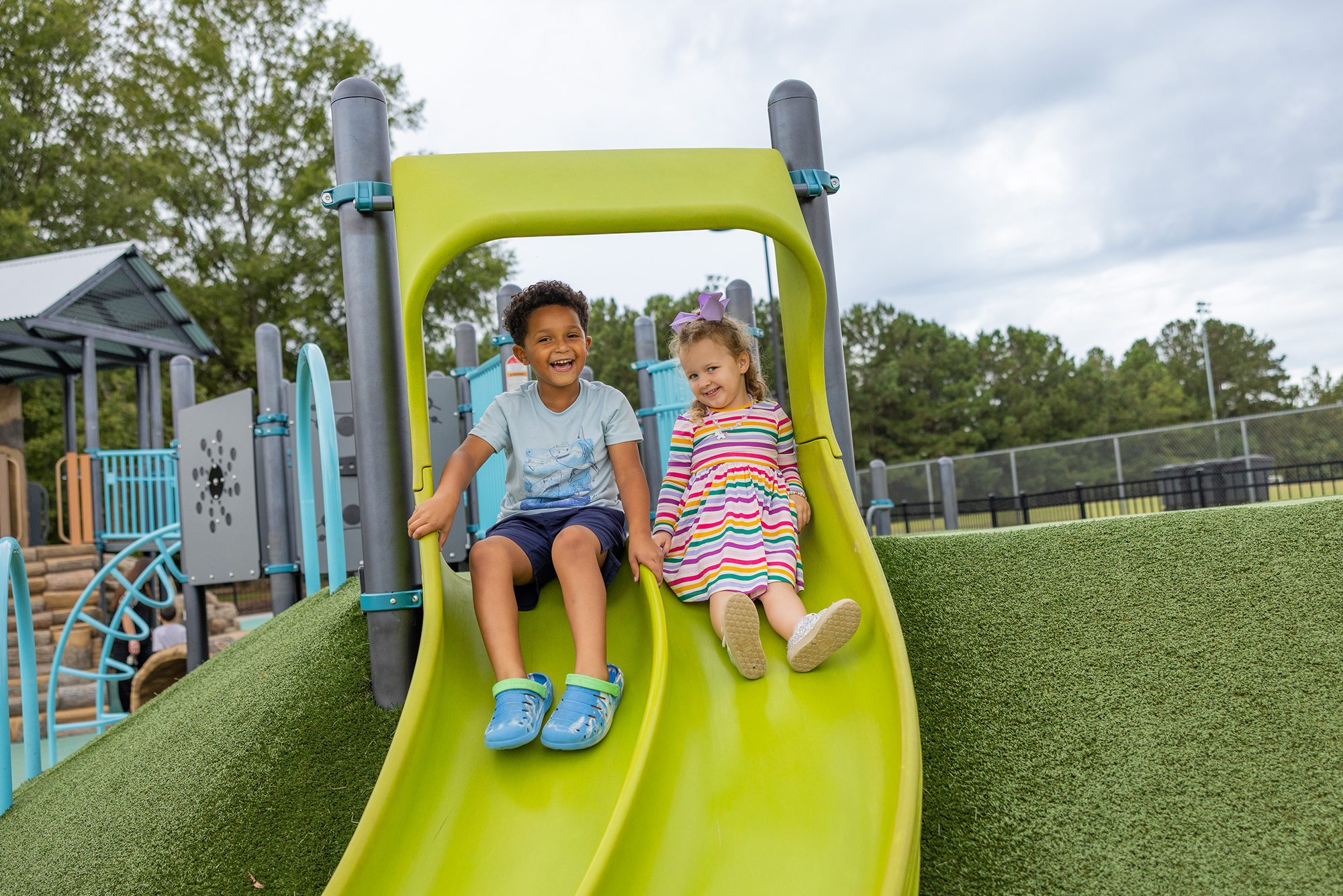 Two smiling children slide down a bright green playground slide outdoors.