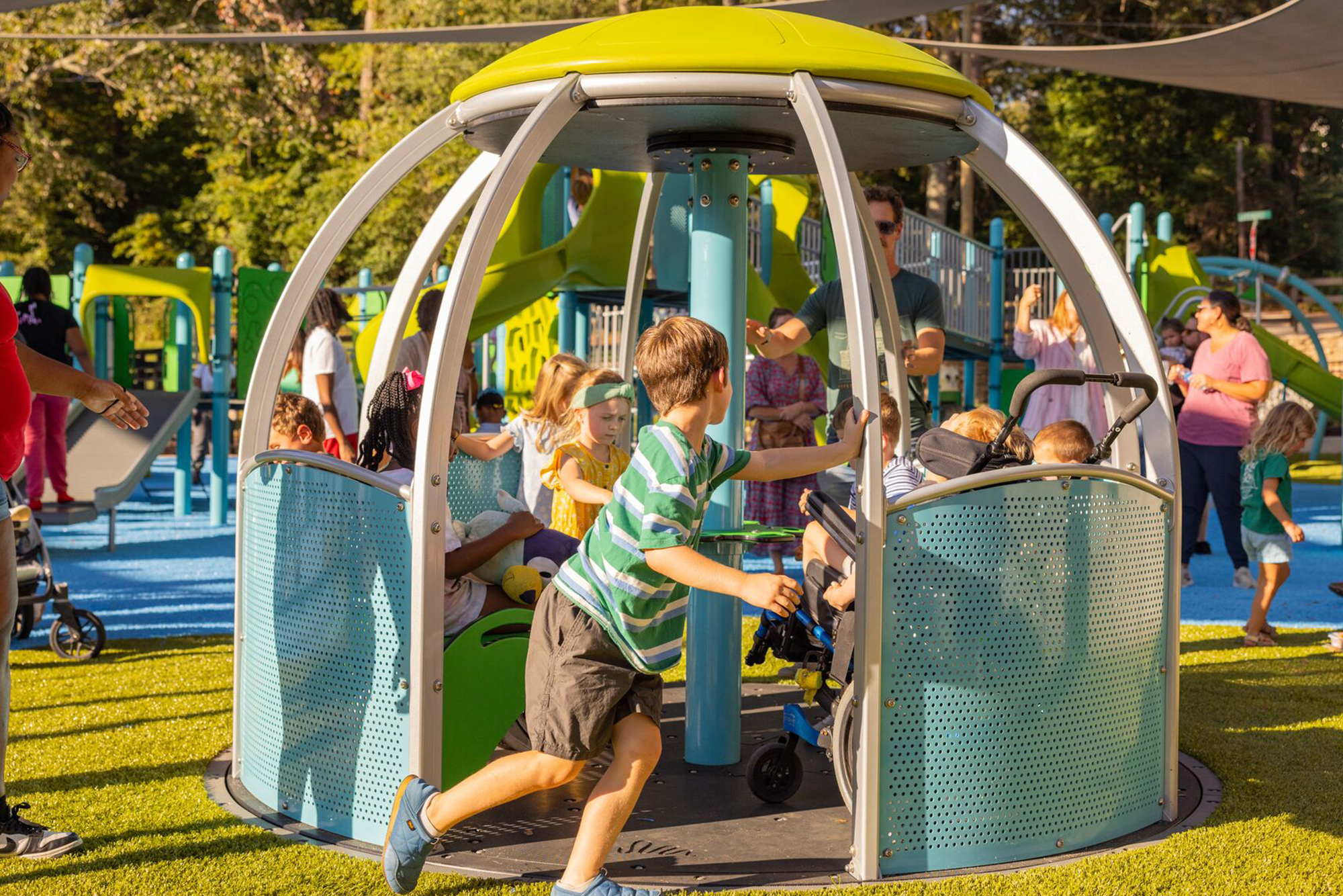 Kids on an inclusive merry-go-round at a sunny playground as adults watch nearby.