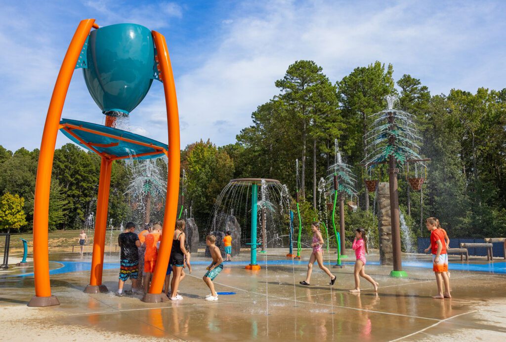 Kids run and splash through water jets at an outdoor splash pad under a bright, sunny sky.