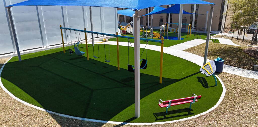 Swings, slides, benches, and shaded areas on artificial grass by a building in a playground.