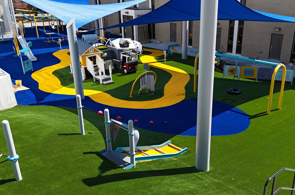 Playground with blue and yellow rubber flooring, various play structures, and large shade sails overhead.