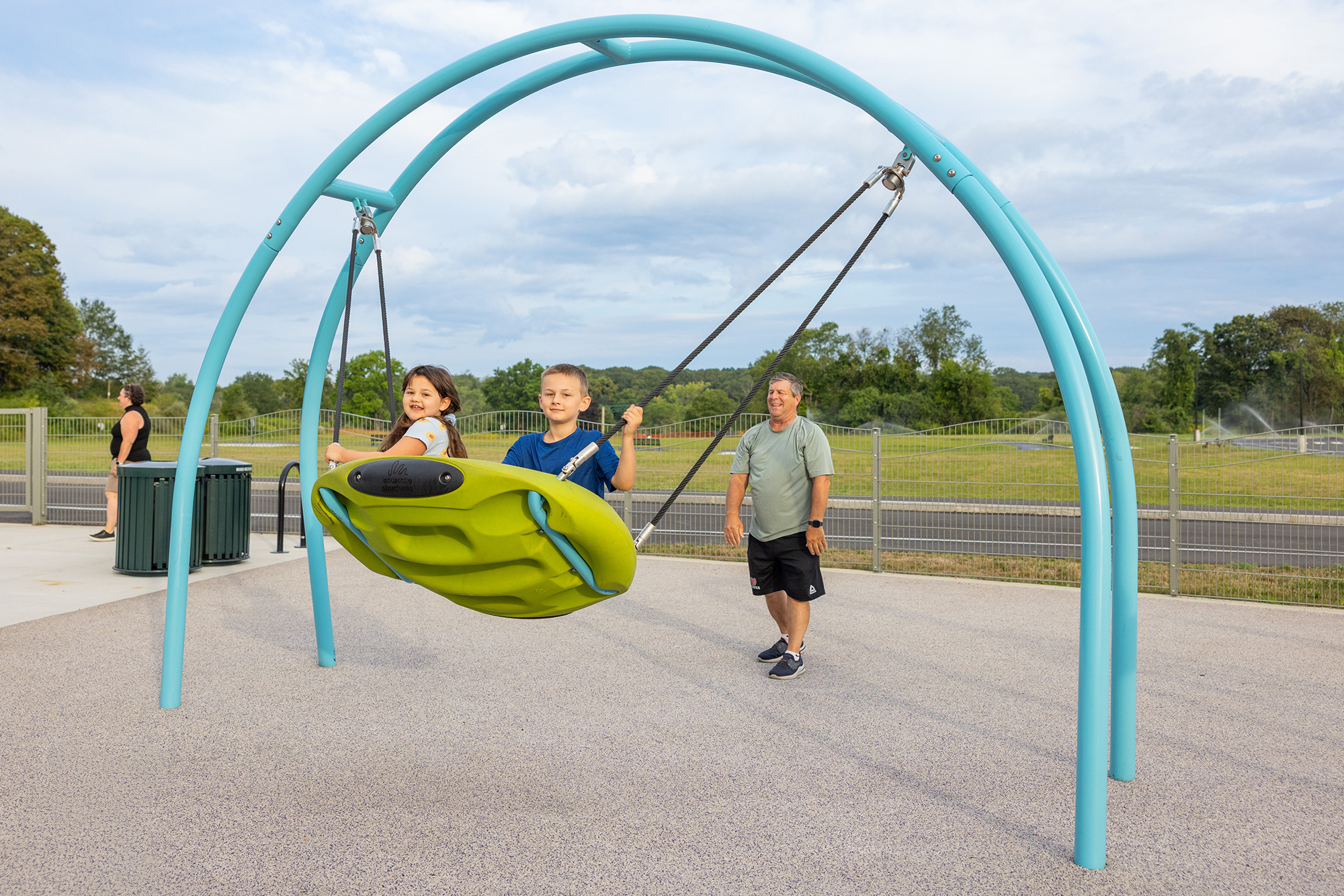 An adult man pushes two children on a green swing at a playground under bright sunshine.