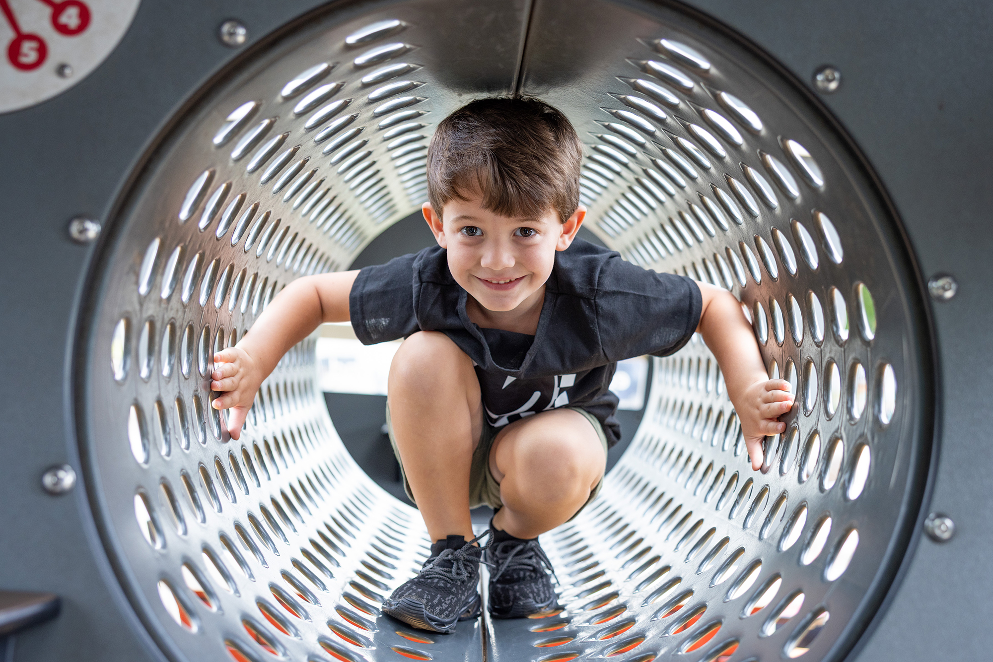 A smiling boy crouches in a metal tunnel at a playground, looking straight at the camera.