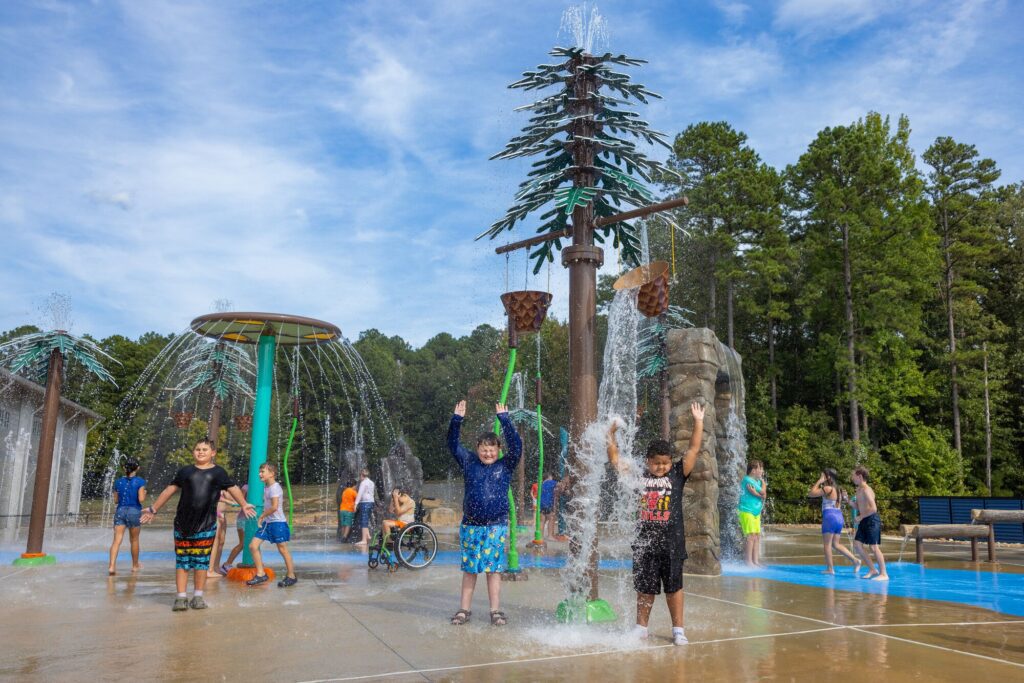 Kids splash and play at an outdoor water park surrounded by fountains and giant tree-like structures.