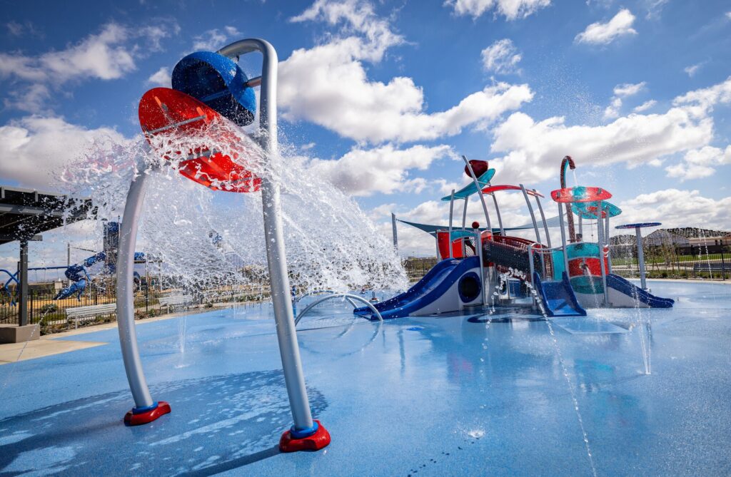 Splash pad with colorful buckets dumping water and slides, set beneath a bright blue sky with scattered white clouds.