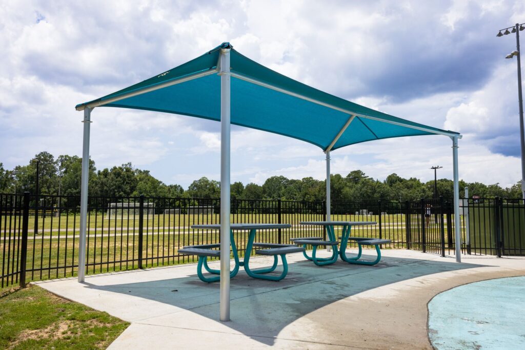 Teal tables and benches in a shaded picnic area beside a sports field on a partly cloudy day.