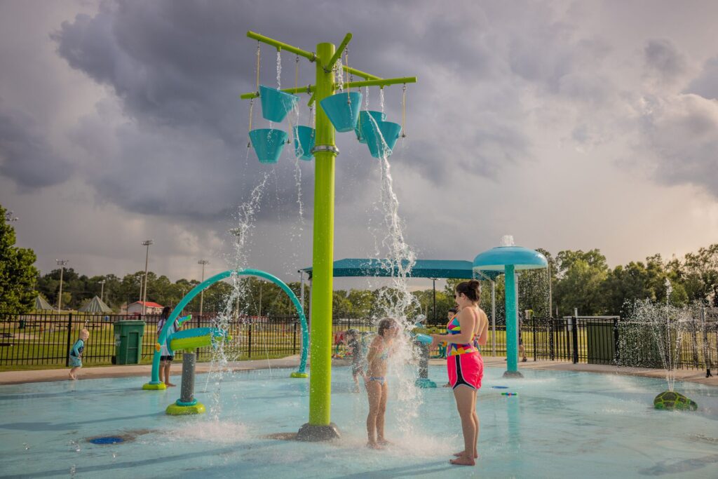 A woman and two children play as water buckets tip over at an outdoor splash pad on a cloudy day.