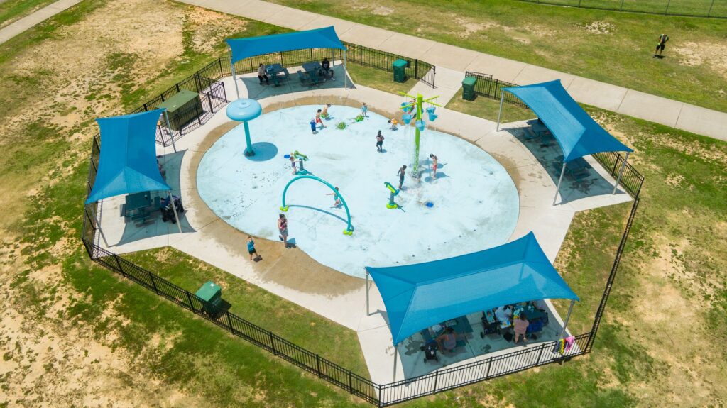 Aerial view of children playing on a splash pad with blue shade structures covering nearby seating areas.