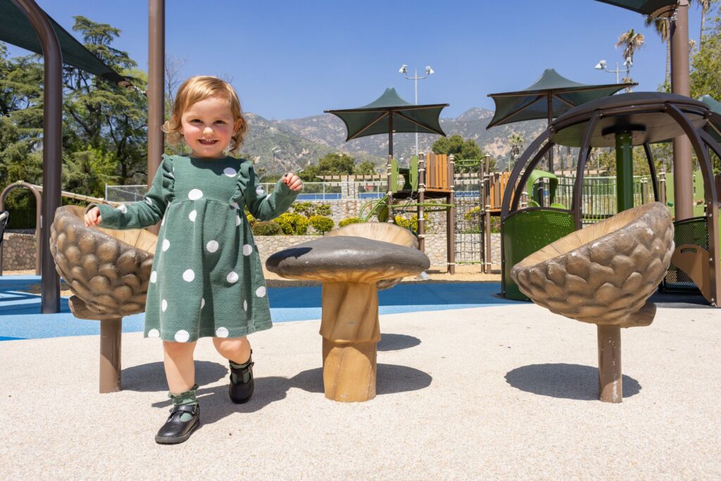 A girl in a green dress stands beside mushroom-shaped seats at an outdoor playground on a sunny day.
