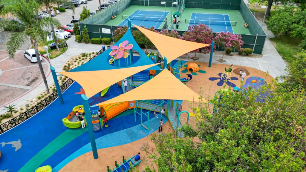 Aerial view of a colorful playground with shade sails, children playing, and tennis courts visible in the background.