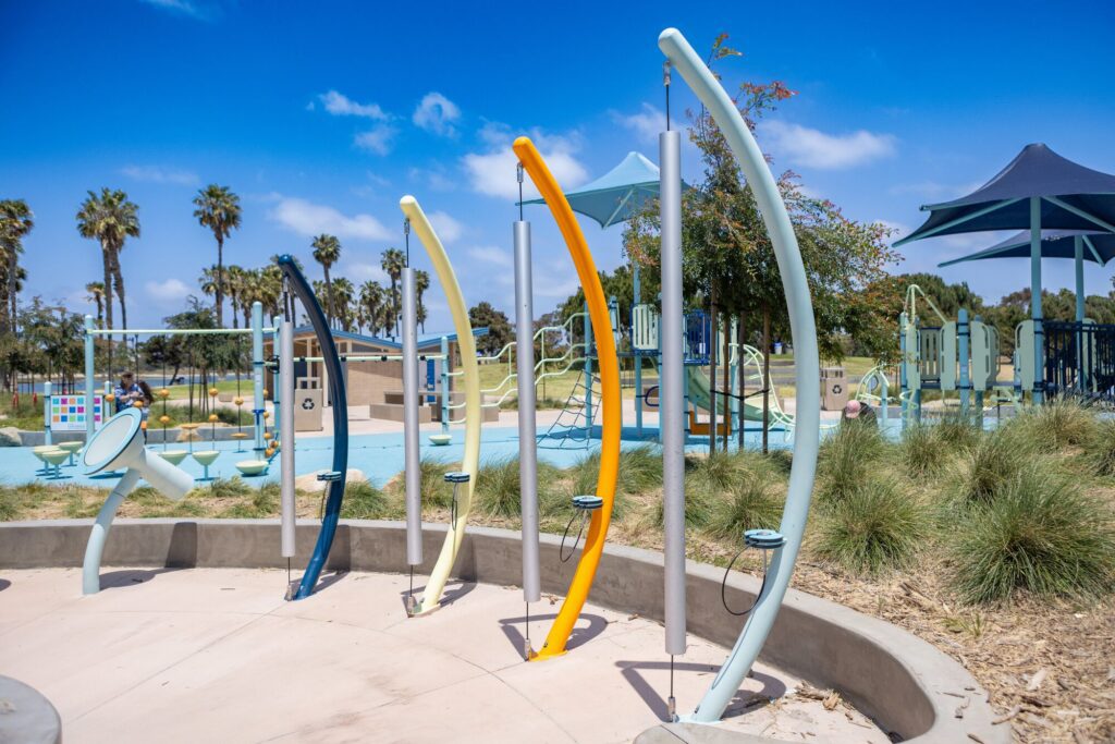 Four bright musical chimes in a sunny playground with green grass and palm trees in the background.