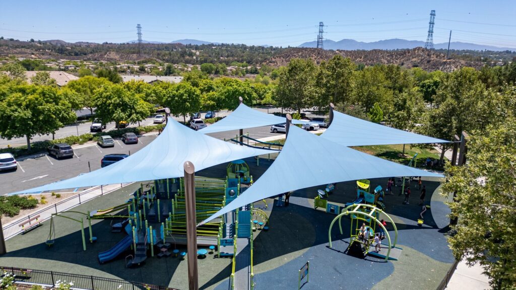 Playground with blue shade sails, bordered by trees and a parking lot, viewed from overhead.