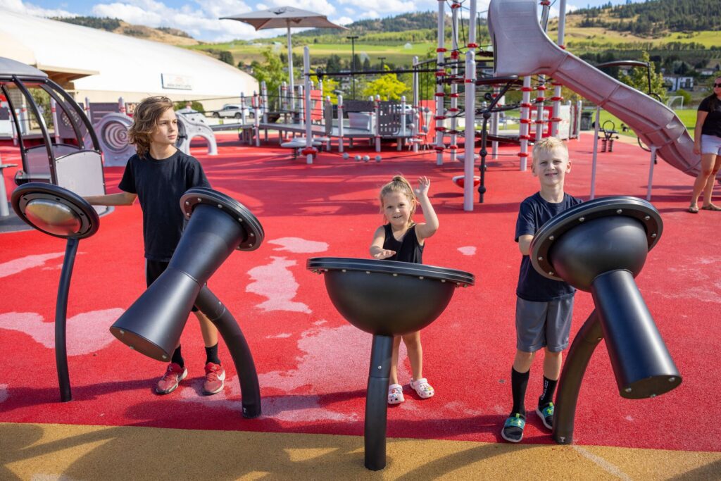 Three kids play outdoor musical instruments at a bright, colorful playground on a sunny day.