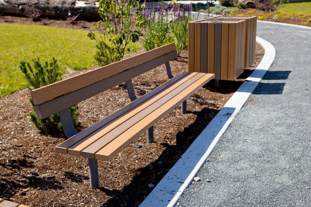 Wooden bench and matching trash bins next to a pathway in a landscaped park on a bright, sunny day.