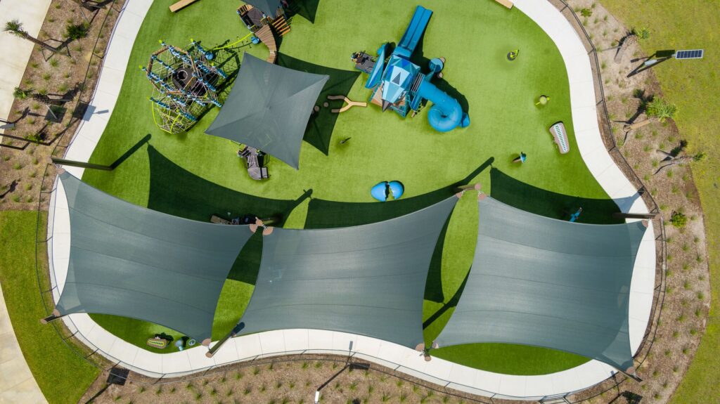Aerial view of a playground featuring green turf, blue play equipment, and shade sails covering the walking paths.