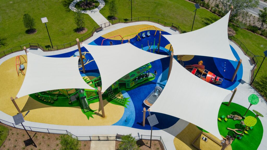 Aerial view of a playground featuring colorful equipment and white shade sails above blue and yellow soft flooring.