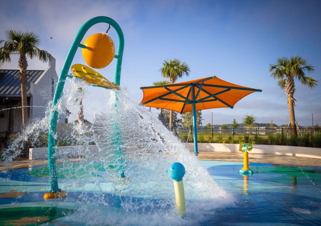 Splash pad with colorful water sprays, tall palm trees, and a big orange umbrella beneath a clear blue sky.
