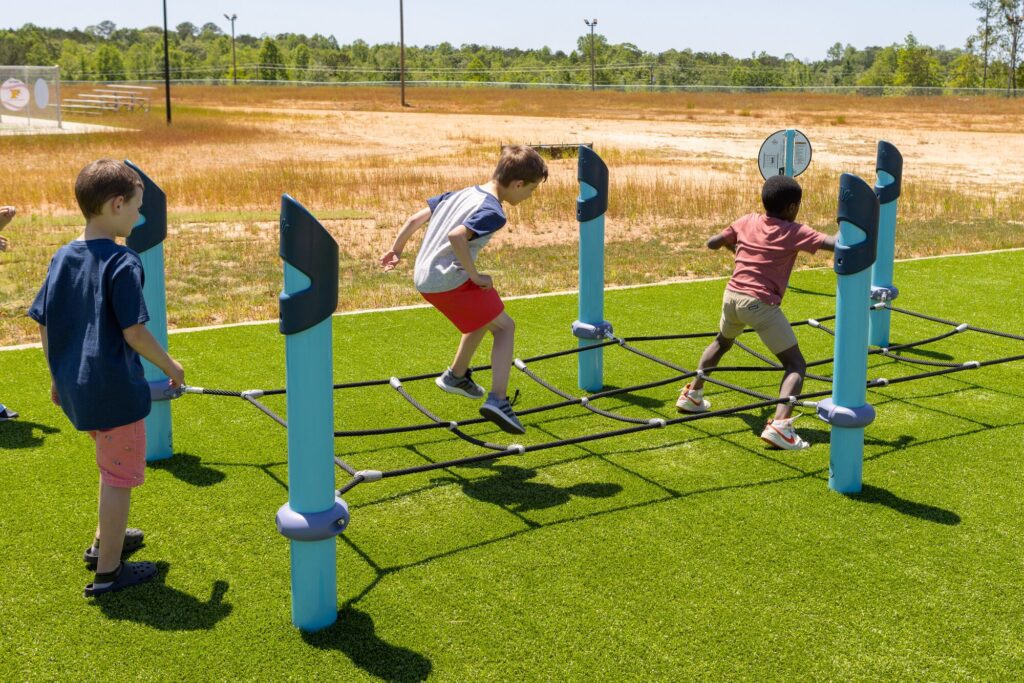 Three boys climb and play on a rope bridge at an outdoor playground under bright sunlight.