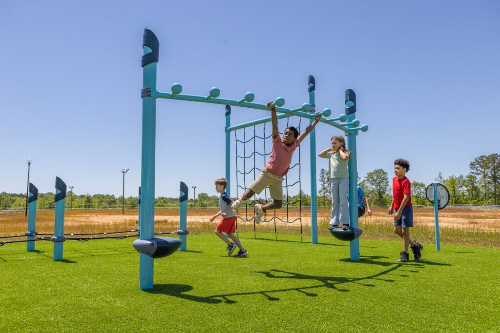 Four kids play on a blue playground set on green turf beneath a clear, sunny sky.
