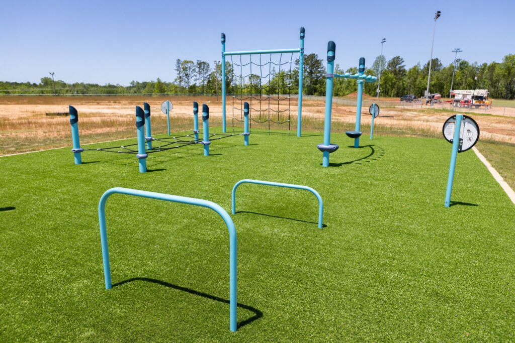 Blue exercise equipment on artificial turf at an outdoor fitness park, bordered by grass and trees.